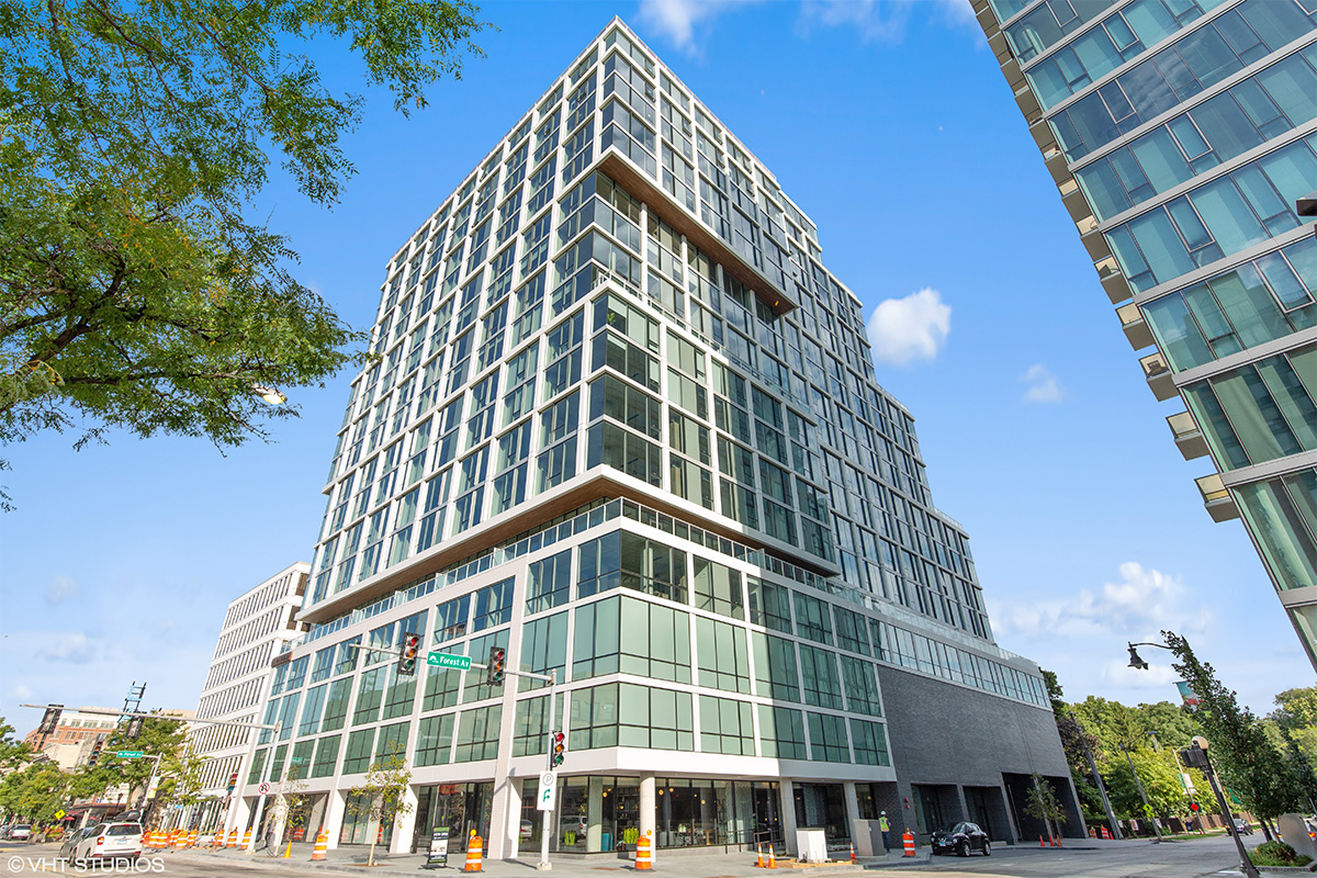 Willton Modern glass and steel building on a city corner with large windows and several stepped levels; blue sky and trees in the background, and traffic cones visible on the street.