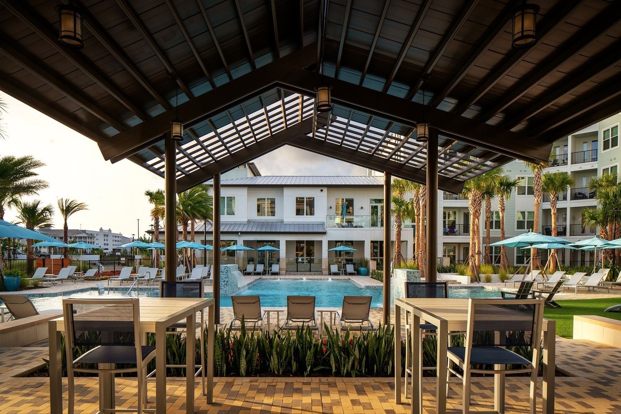 Willton A covered patio with tables and chairs overlooks a resort-style swimming pool surrounded by lounge chairs, palm trees, and umbrella-covered tables, with modern apartment buildings in the background.